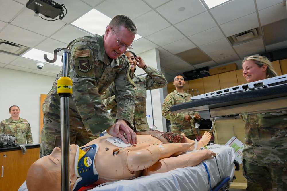 Military personnel in U.S. Army/Air Force OCP (Operational Camouflage Pattern) uniforms are engaged in a medical simulation training exercise. A senior service member is examining a full-body medical training mannequin on a gurney, which appears to have simulated wounds or medical attachments. The setting is an indoor medical simulation laboratory with multiple personnel observing and participating.