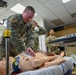 Military personnel in U.S. Army/Air Force OCP (Operational Camouflage Pattern) uniforms are engaged in a medical simulation training exercise. A senior service member is examining a full-body medical training mannequin on a gurney, which appears to have simulated wounds or medical attachments. The setting is an indoor medical simulation laboratory with multiple personnel observing and participating.
