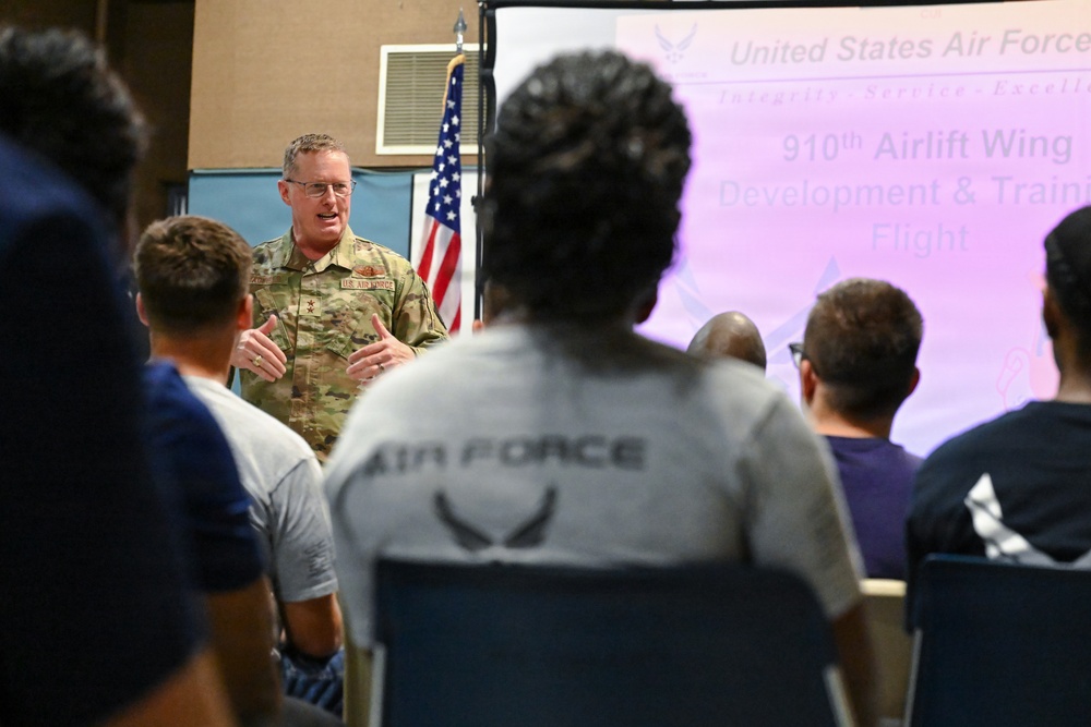 A U.S. Air Force officer in OCP (Operational Camouflage Pattern) uniform is addressing a group of personnel in a briefing or orientation setting. A presentation screen behind him displays '910th Airlift Wing Development &amp; Training Flight' with the United States Air Force logo and motto. The audience, some wearing Air Force PT gear, is seated facing the speaker.