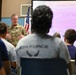 A U.S. Air Force officer in OCP (Operational Camouflage Pattern) uniform is addressing a group of personnel in a briefing or orientation setting. A presentation screen behind him displays '910th Airlift Wing Development &amp; Training Flight' with the United States Air Force logo and motto. The audience, some wearing Air Force PT gear, is seated facing the speaker.