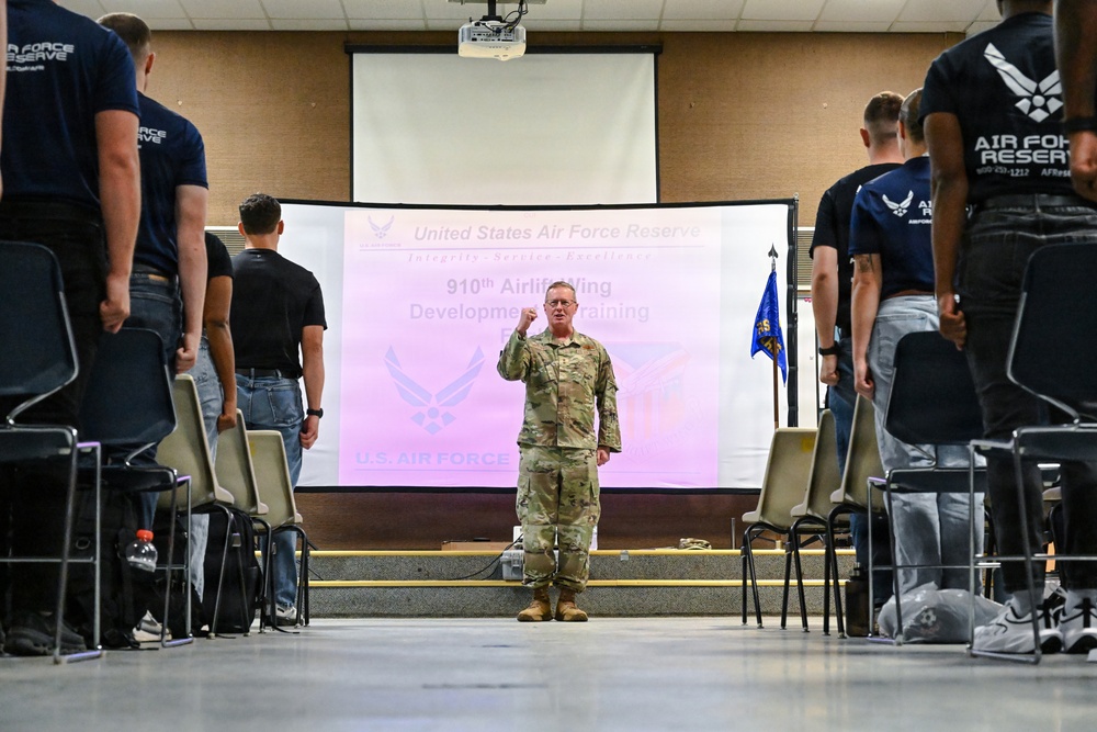 A uniformed U.S. Air Force Reserve officer stands at the front of a briefing room, addressing a group of personnel standing in rows on either side of a center aisle. The presentation screen behind him displays a slide for the '910th Airlift Wing Development Training' program under the United States Air Force Reserve. The setting appears to be a formal training or orientation session.