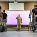 A uniformed U.S. Air Force Reserve officer stands at the front of a briefing room, addressing a group of personnel standing in rows on either side of a center aisle. The presentation screen behind him displays a slide for the '910th Airlift Wing Development Training' program under the United States Air Force Reserve. The setting appears to be a formal training or orientation session.