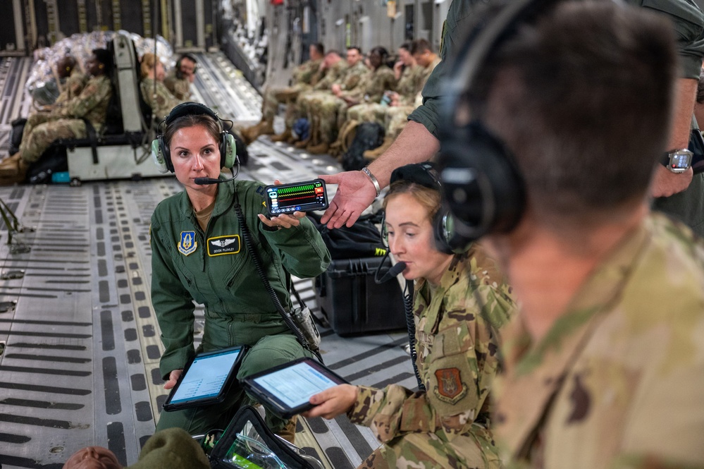 Military personnel are conducting what appears to be an aeromedical evacuation exercise or operation inside a large military transport aircraft, likely a C-17 Globemaster III. A female service member in green flight suit with flight nurse wings is being shown a medical monitoring device displaying vital signs on a smartphone screen. Multiple personnel in both flight suits and combat uniforms are using tablets and headsets, suggesting a coordinated medical or training operation.