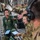 Military personnel are conducting what appears to be an aeromedical evacuation exercise or operation inside a large military transport aircraft, likely a C-17 Globemaster III. A female service member in green flight suit with flight nurse wings is being shown a medical monitoring device displaying vital signs on a smartphone screen. Multiple personnel in both flight suits and combat uniforms are using tablets and headsets, suggesting a coordinated medical or training operation.