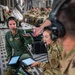 Military personnel are conducting what appears to be an aeromedical evacuation exercise or operation inside a large military transport aircraft, likely a C-17 Globemaster III. A female service member in green flight suit with flight nurse wings is being shown a medical monitoring device displaying vital signs on a smartphone screen. Multiple personnel in both flight suits and combat uniforms are using tablets and headsets, suggesting a coordinated medical or training operation.