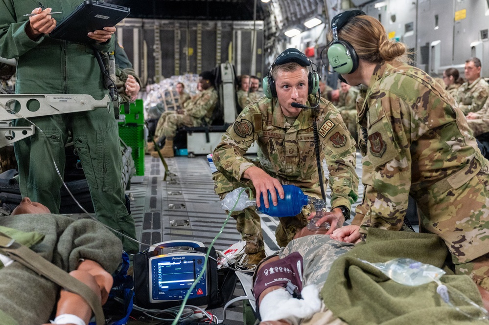 Military medical personnel in OCP (Operational Camouflage Pattern) uniforms are conducting aeromedical evacuation procedures aboard a large military transport aircraft, likely a C-17 Globemaster III. Two medics wearing aviation headsets are attending to patients on litters, using a bag-valve mask and monitoring equipment. The scene appears to be either a training exercise or an actual aeromedical evacuation mission, with numerous additional personnel seated in the background.
