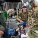 Military medical personnel in OCP (Operational Camouflage Pattern) uniforms are conducting aeromedical evacuation procedures aboard a large military transport aircraft, likely a C-17 Globemaster III. Two medics wearing aviation headsets are attending to patients on litters, using a bag-valve mask and monitoring equipment. The scene appears to be either a training exercise or an actual aeromedical evacuation mission, with numerous additional personnel seated in the background.