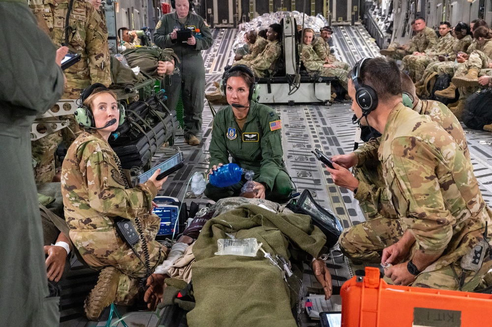 Military medical personnel are conducting an aeromedical evacuation exercise or operation inside what appears to be a C-17 Globemaster III military transport aircraft. A team of three primary medical providers — two in camouflage uniforms and one in a green flight suit with a US flag patch — are attending to a patient on a litter with medical equipment. Additional military personnel are seated along the aircraft walls in the background.