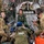 Military medical personnel are conducting an aeromedical evacuation exercise or operation inside what appears to be a C-17 Globemaster III military transport aircraft. A team of three primary medical providers — two in camouflage uniforms and one in a green flight suit with a US flag patch — are attending to a patient on a litter with medical equipment. Additional military personnel are seated along the aircraft walls in the background.