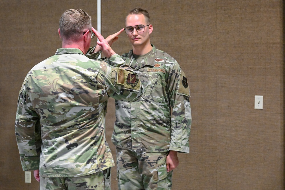 Two U.S. Air Force personnel in OCP (Operational Camouflage Pattern) uniforms are engaged in a formal military salute exchange, likely during a ceremony or official event. The individual facing the camera wears a 'U.S. Air Force' tape and what appears to be aviator wings on their uniform. The setting appears to be an indoor venue with wood-paneled walls, consistent with a conference room or event hall.