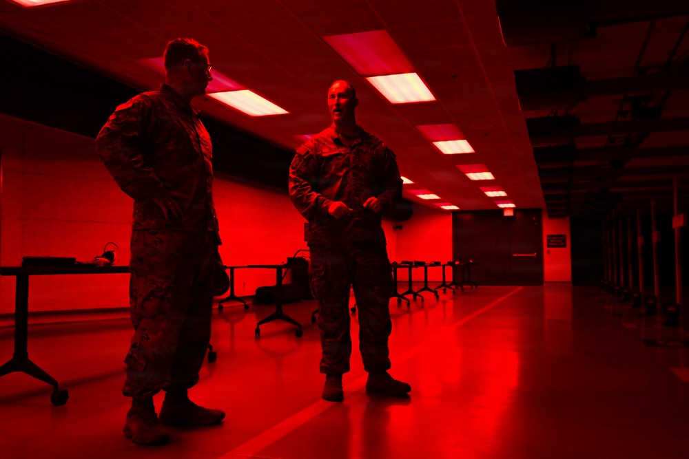 Two military personnel in camouflage uniforms are standing and conversing inside a large room illuminated by red lighting. The space appears to be a briefing room or operations center with rows of workstations and chairs visible in the background. The red ambient lighting dominates the entire scene, casting a dramatic tone throughout the facility.