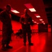 Two military personnel in camouflage uniforms are standing and conversing inside a large room illuminated by red lighting. The space appears to be a briefing room or operations center with rows of workstations and chairs visible in the background. The red ambient lighting dominates the entire scene, casting a dramatic tone throughout the facility.