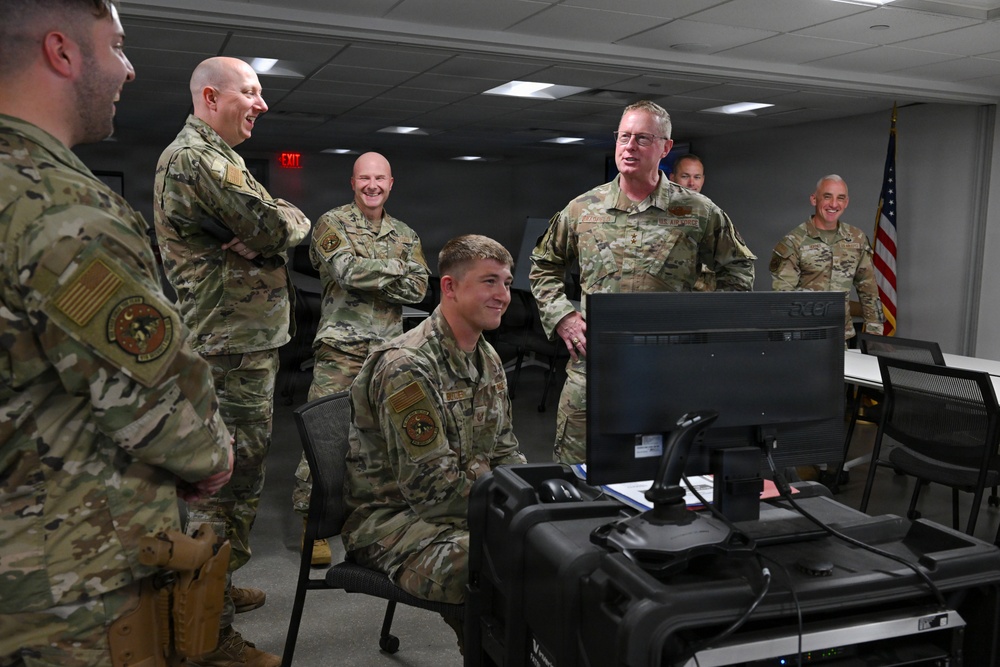 A group of military personnel in OCP (Operational Camouflage Pattern) uniforms are gathered in what appears to be a command or operations room. One service member is seated at a workstation with a computer monitor and what appears to be a control console, while several others stand around engaging in conversation. The atmosphere appears relaxed and informal, with multiple individuals smiling.