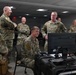 A group of military personnel in OCP (Operational Camouflage Pattern) uniforms are gathered in what appears to be a command or operations room. One service member is seated at a workstation with a computer monitor and what appears to be a control console, while several others stand around engaging in conversation. The atmosphere appears relaxed and informal, with multiple individuals smiling.