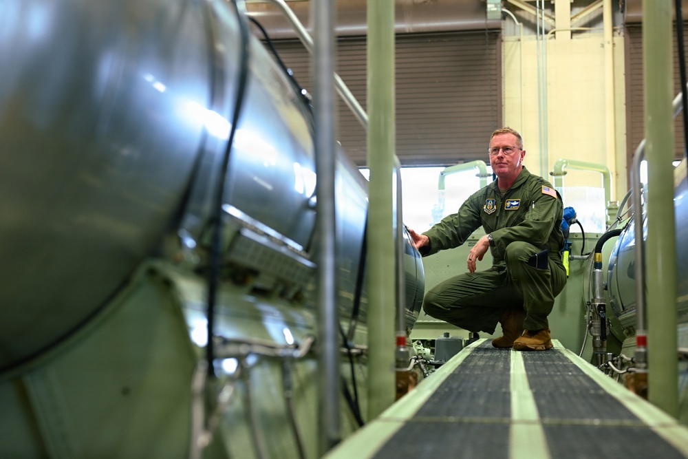 A military personnel member wearing an olive green flight suit with US Air Force patches is crouching between large cylindrical metallic equipment in what appears to be an industrial or aerospace facility. The individual appears to be inspecting or examining the equipment. The setting suggests a maintenance bay, test facility, or storage area for large aerospace or military hardware.