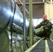 A military personnel member wearing an olive green flight suit with US Air Force patches is crouching between large cylindrical metallic equipment in what appears to be an industrial or aerospace facility. The individual appears to be inspecting or examining the equipment. The setting suggests a maintenance bay, test facility, or storage area for large aerospace or military hardware.