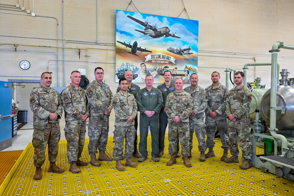 A group of approximately eleven military personnel pose for a formal group photo inside an industrial facility. They are standing on yellow safety grating in front of a large painted mural depicting aircraft and the text 'DOD'S ONE AND ONLY AERIAL SPRAY MAINTENANCE'. The group includes individuals in camouflage uniforms and one person in a green flight suit.