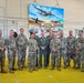 A group of approximately eleven military personnel pose for a formal group photo inside an industrial facility. They are standing on yellow safety grating in front of a large painted mural depicting aircraft and the text 'DOD'S ONE AND ONLY AERIAL SPRAY MAINTENANCE'. The group includes individuals in camouflage uniforms and one person in a green flight suit.