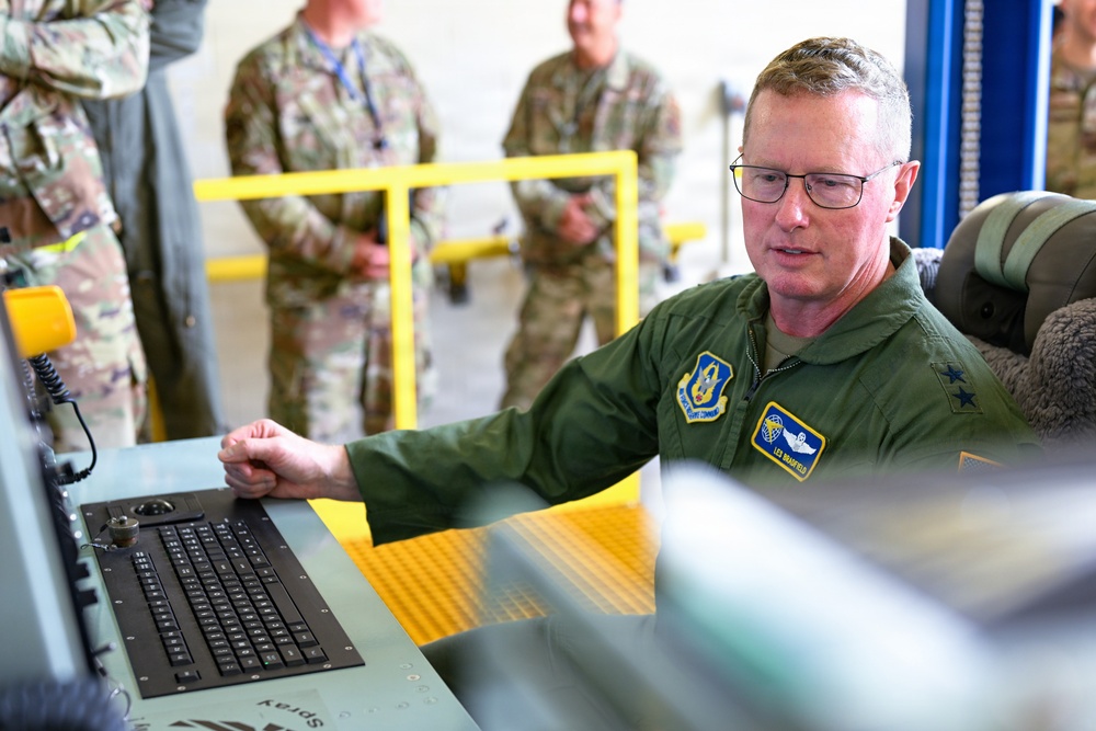 A senior military officer in a green flight suit is seated at a technical workstation with a keyboard and control equipment, appearing to operate or demonstrate the system. Several other military personnel in camouflage uniforms observe in the background. The setting appears to be a military facility or hangar with yellow safety railings visible.