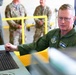 A senior military officer in a green flight suit is seated at a technical workstation with a keyboard and control equipment, appearing to operate or demonstrate the system. Several other military personnel in camouflage uniforms observe in the background. The setting appears to be a military facility or hangar with yellow safety railings visible.