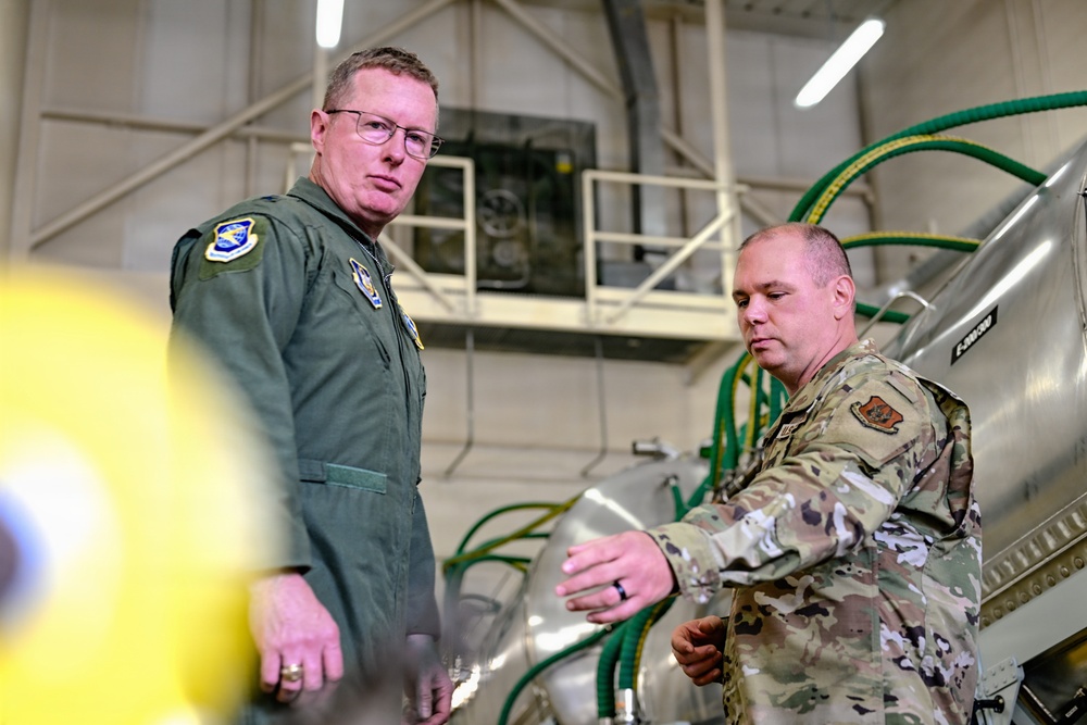 Two U.S. Air Force personnel are inside what appears to be a military hangar or maintenance facility. One individual is wearing an olive green flight suit with patches, while the other is in OCP (Operational Camouflage Pattern) uniform. They appear to be inspecting or discussing equipment, likely munitions or aircraft-related hardware.