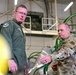 Two U.S. Air Force personnel are inside what appears to be a military hangar or maintenance facility. One individual is wearing an olive green flight suit with patches, while the other is in OCP (Operational Camouflage Pattern) uniform. They appear to be inspecting or discussing equipment, likely munitions or aircraft-related hardware.
