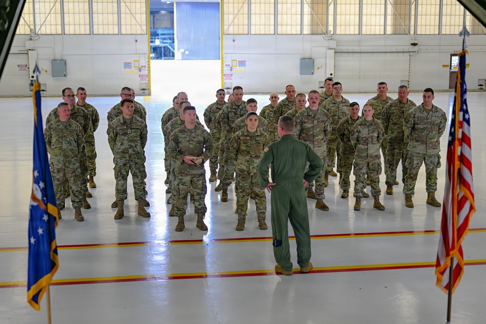 A military formation is taking place inside an aircraft hangar, where a single officer in a green flight suit stands with hands on hips addressing a group of approximately 25-30 soldiers in OCP (Operational Camouflage Pattern) uniforms. Two flags are visible flanking the scene — the United States flag on the right and what appears to be an Air Force flag on the left. The setting, uniforms, and formal arrangement suggest an official military briefing, ceremony, or unit address.