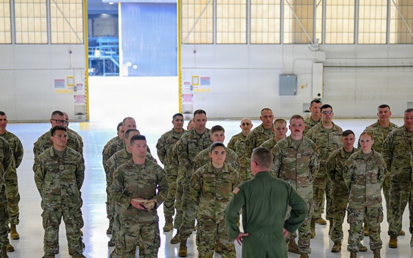 A military formation is taking place inside an aircraft hangar, where a single officer in a green flight suit stands with hands on hips addressing a group of approximately 25-30 soldiers in OCP (Operational Camouflage Pattern) uniforms. Two flags are visible flanking the scene — the United States flag on the right and what appears to be an Air Force flag on the left. The setting, uniforms, and formal arrangement suggest an official military briefing, ceremony, or unit address.