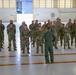 A military formation is taking place inside an aircraft hangar, where a single officer in a green flight suit stands with hands on hips addressing a group of approximately 25-30 soldiers in OCP (Operational Camouflage Pattern) uniforms. Two flags are visible flanking the scene — the United States flag on the right and what appears to be an Air Force flag on the left. The setting, uniforms, and formal arrangement suggest an official military briefing, ceremony, or unit address.