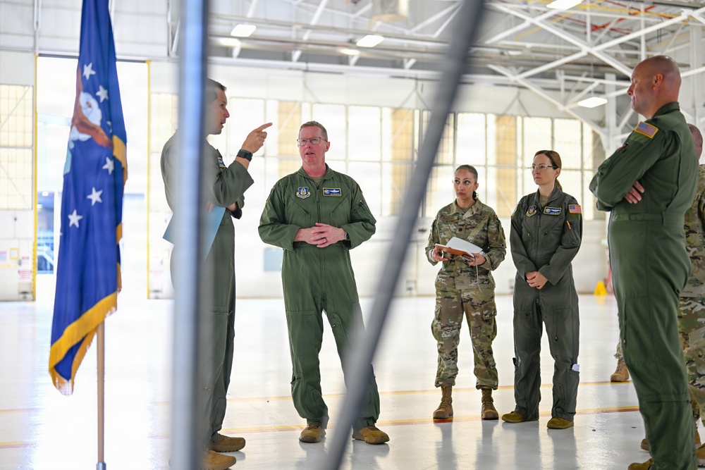 A group of U.S. Air Force personnel are gathered inside a large aircraft hangar for what appears to be a briefing or discussion. One individual in a flight suit is gesturing and speaking, while others listen attentively. A U.S. Air Force flag with gold fringe is visible on the left side of the frame.