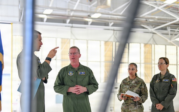 A group of U.S. Air Force personnel are gathered inside a large aircraft hangar for what appears to be a briefing or discussion. One individual in a flight suit is gesturing and speaking, while others listen attentively. A U.S. Air Force flag with gold fringe is visible on the left side of the frame.