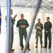 A group of U.S. Air Force personnel are gathered inside a large aircraft hangar for what appears to be a briefing or discussion. One individual in a flight suit is gesturing and speaking, while others listen attentively. A U.S. Air Force flag with gold fringe is visible on the left side of the frame.