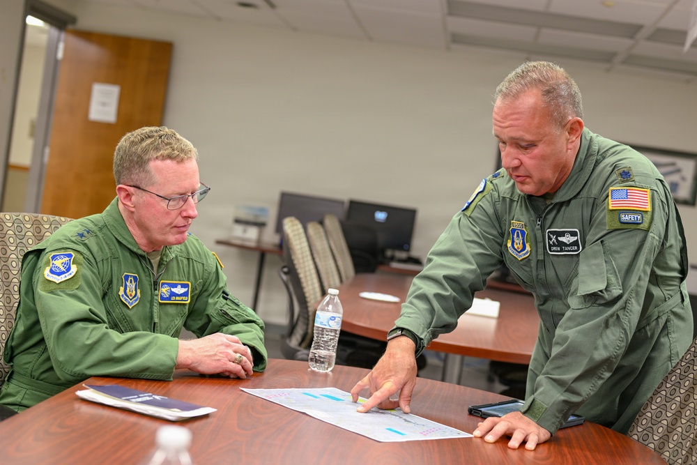 Two U.S. Air Force personnel in green flight suits are seated and standing at a conference table, reviewing what appears to be a map or chart document. The individual on the right, whose name tag reads 'DREW TANCER,' is pointing to a specific area on the document while the other individual, whose name tag reads 'LEN MANSFIELD,' observes attentively. Both wear unit patches, pilot wings, and American flag insignia consistent with U.S. Air Force flight crew uniforms.