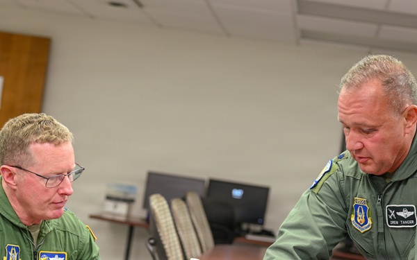 Two U.S. Air Force personnel in green flight suits are seated and standing at a conference table, reviewing what appears to be a map or chart document. The individual on the right, whose name tag reads 'DREW TANCER,' is pointing to a specific area on the document while the other individual, whose name tag reads 'LEN MANSFIELD,' observes attentively. Both wear unit patches, pilot wings, and American flag insignia consistent with U.S. Air Force flight crew uniforms.