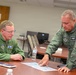 Two U.S. Air Force personnel in green flight suits are seated and standing at a conference table, reviewing what appears to be a map or chart document. The individual on the right, whose name tag reads 'DREW TANCER,' is pointing to a specific area on the document while the other individual, whose name tag reads 'LEN MANSFIELD,' observes attentively. Both wear unit patches, pilot wings, and American flag insignia consistent with U.S. Air Force flight crew uniforms.