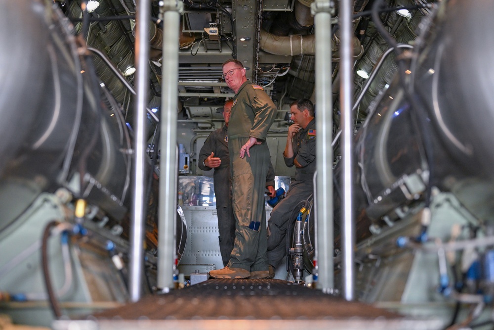 Three individuals wearing olive/sage green military flight suits are gathered inside what appears to be the cargo hold or interior of a large military aircraft. The image is taken from a low angle through vertical support struts, revealing complex mechanical infrastructure including ducting, wiring, and hydraulic lines overhead. The personnel appear to be engaged in discussion or inspection activities.