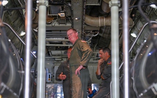 Three individuals wearing olive/sage green military flight suits are gathered inside what appears to be the cargo hold or interior of a large military aircraft. The image is taken from a low angle through vertical support struts, revealing complex mechanical infrastructure including ducting, wiring, and hydraulic lines overhead. The personnel appear to be engaged in discussion or inspection activities.