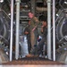 Three individuals wearing olive/sage green military flight suits are gathered inside what appears to be the cargo hold or interior of a large military aircraft. The image is taken from a low angle through vertical support struts, revealing complex mechanical infrastructure including ducting, wiring, and hydraulic lines overhead. The personnel appear to be engaged in discussion or inspection activities.