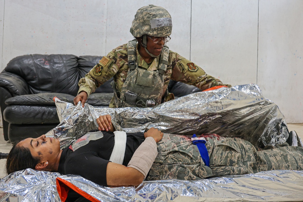A U.S. Air Force service member in full combat gear and ACH helmet is providing medical care to a casualty lying on a litter covered with a mylar emergency blanket. The casualty has visible bandaging on the neck/shoulder area and simulated injuries including moulage makeup. This appears to be a military medical training exercise or readiness drill.