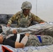 A U.S. Air Force service member in full combat gear and ACH helmet is providing medical care to a casualty lying on a litter covered with a mylar emergency blanket. The casualty has visible bandaging on the neck/shoulder area and simulated injuries including moulage makeup. This appears to be a military medical training exercise or readiness drill.