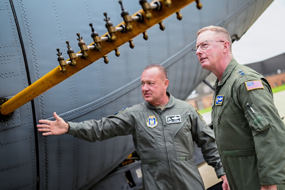 Two military personnel in olive green flight suits are examining the exterior of a large military aircraft. One individual is gesturing toward a yellow fuel or hydraulic manifold bar with multiple valve fittings attached to the aircraft's fuselage. The person on the right displays two stars on their collar, indicating a Major General rank, and wears a U.S. flag patch on their sleeve.