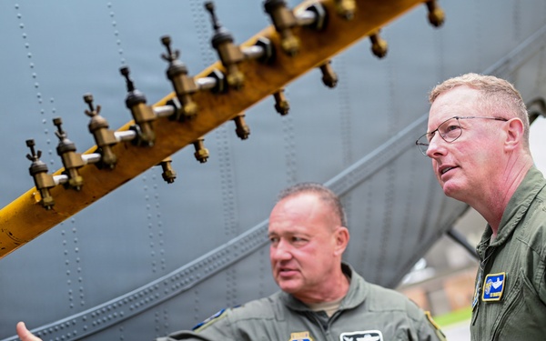 Two military personnel in olive green flight suits are examining the exterior of a large military aircraft. One individual is gesturing toward a yellow fuel or hydraulic manifold bar with multiple valve fittings attached to the aircraft's fuselage. The person on the right displays two stars on their collar, indicating a Major General rank, and wears a U.S. flag patch on their sleeve.