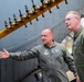 Two military personnel in olive green flight suits are examining the exterior of a large military aircraft. One individual is gesturing toward a yellow fuel or hydraulic manifold bar with multiple valve fittings attached to the aircraft's fuselage. The person on the right displays two stars on their collar, indicating a Major General rank, and wears a U.S. flag patch on their sleeve.