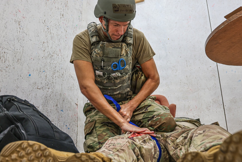 A military personnel wearing a combat helmet and tactical plate carrier vest is administering first aid or medical treatment to a casualty on the ground. The individual appears to be applying pressure or a tourniquet to a simulated wound on the casualty's leg, suggesting this may be a combat casualty care training exercise. Blue medical scissors are visibly clipped to the front of the vest.