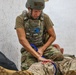 A military personnel wearing a combat helmet and tactical plate carrier vest is administering first aid or medical treatment to a casualty on the ground. The individual appears to be applying pressure or a tourniquet to a simulated wound on the casualty's leg, suggesting this may be a combat casualty care training exercise. Blue medical scissors are visibly clipped to the front of the vest.