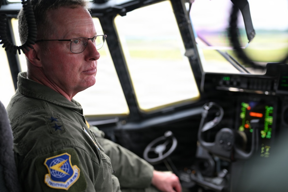 A military aviator in an olive green flight suit is seated in the cockpit of a large military aircraft, likely a C-130 or similar transport aircraft. He is wearing glasses, a communications headset, and displays a patch on his left shoulder consistent with Air Mobility Command (USAF). The cockpit instruments and large forward windows are visible, with an airfield visible in the background.
