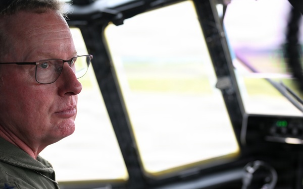 A military aviator in an olive green flight suit is seated in the cockpit of a large military aircraft, likely a C-130 or similar transport aircraft. He is wearing glasses, a communications headset, and displays a patch on his left shoulder consistent with Air Mobility Command (USAF). The cockpit instruments and large forward windows are visible, with an airfield visible in the background.