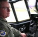 A military aviator in an olive green flight suit is seated in the cockpit of a large military aircraft, likely a C-130 or similar transport aircraft. He is wearing glasses, a communications headset, and displays a patch on his left shoulder consistent with Air Mobility Command (USAF). The cockpit instruments and large forward windows are visible, with an airfield visible in the background.