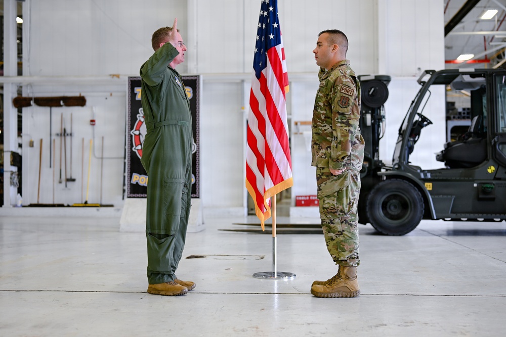 Two U.S. Air Force personnel are participating in what appears to be a formal military ceremony, likely a swearing-in or reenlistment, inside a hangar facility. The officer on the left, wearing an olive green flight suit, has his right hand raised in an oath gesture, while the airman on the right in OCP (Operational Camouflage Pattern) uniform stands at attention. An American flag on a ceremonial stand is positioned between them.