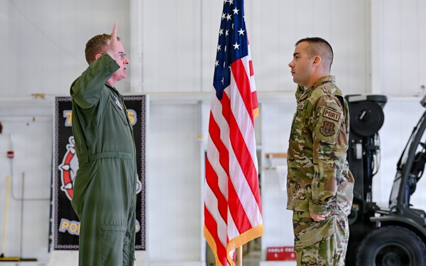 Two U.S. Air Force personnel are participating in what appears to be a formal military ceremony, likely a swearing-in or reenlistment, inside a hangar facility. The officer on the left, wearing an olive green flight suit, has his right hand raised in an oath gesture, while the airman on the right in OCP (Operational Camouflage Pattern) uniform stands at attention. An American flag on a ceremonial stand is positioned between them.